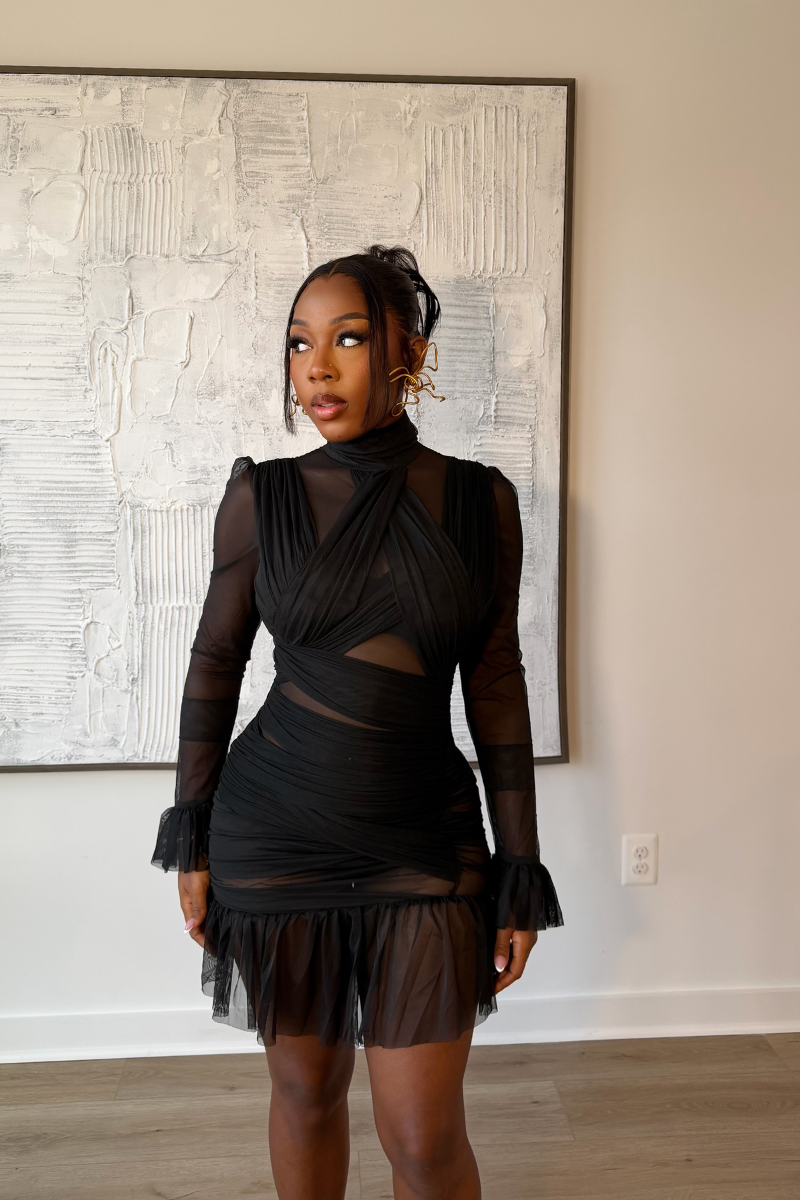 Woman wearing Demuluxe Vesper Wrap Dress, a black mesh mini with draped crisscross bodice and sheer sleeves
