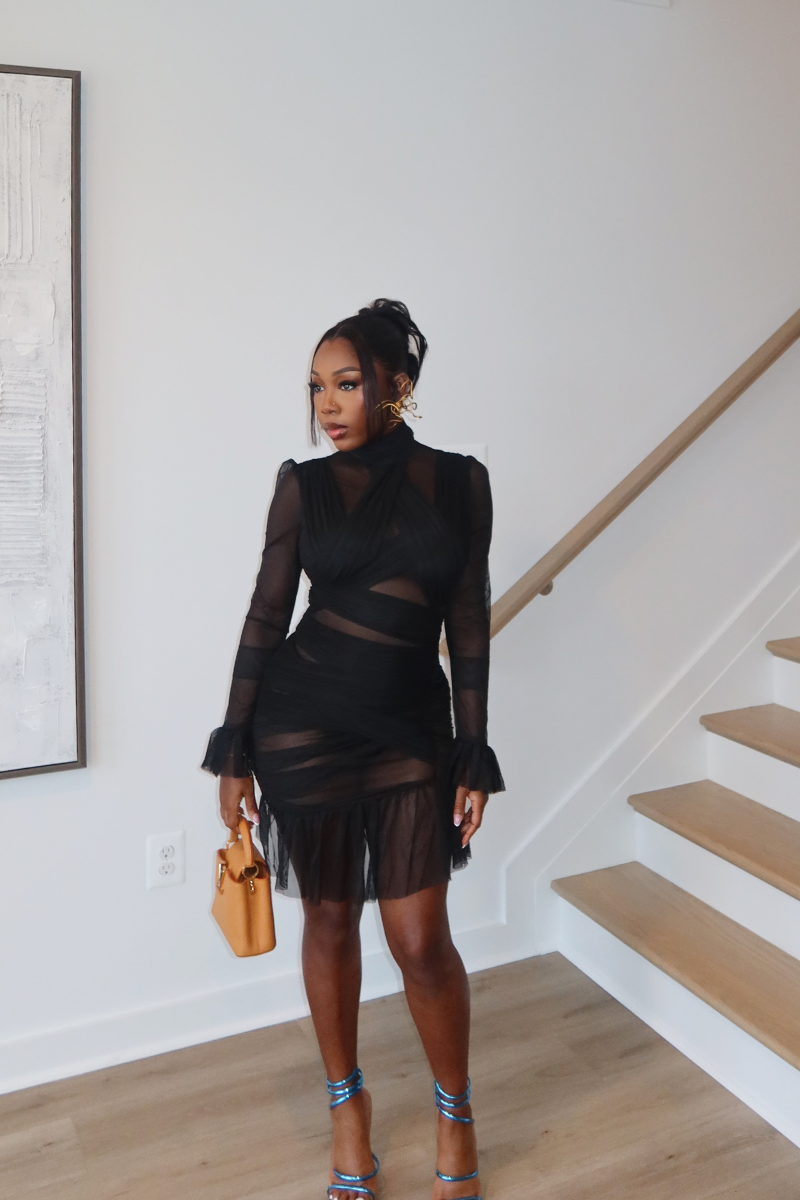 Woman wearing Demuluxe Vesper Wrap Dress, a black mesh mini with draped crisscross bodice and sheer sleeves