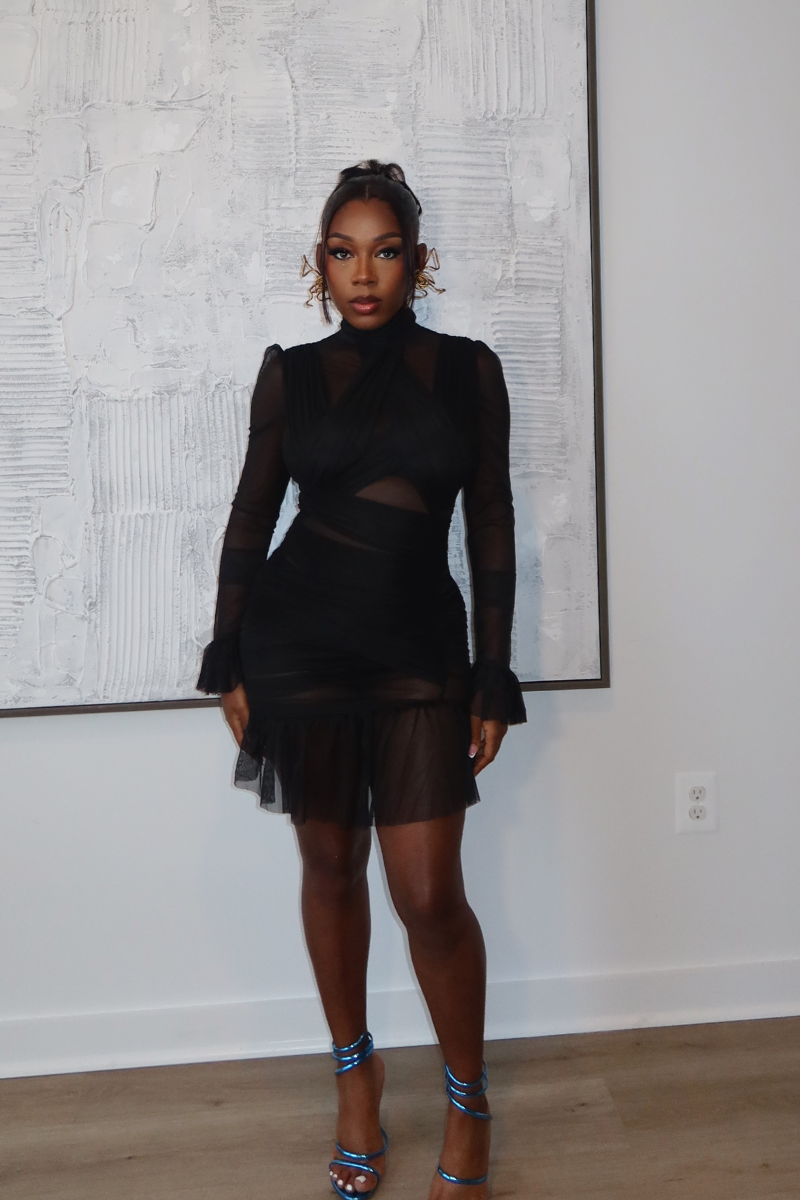 Woman wearing Demuluxe Vesper Wrap Dress, a black mesh mini with draped crisscross bodice and sheer sleeves