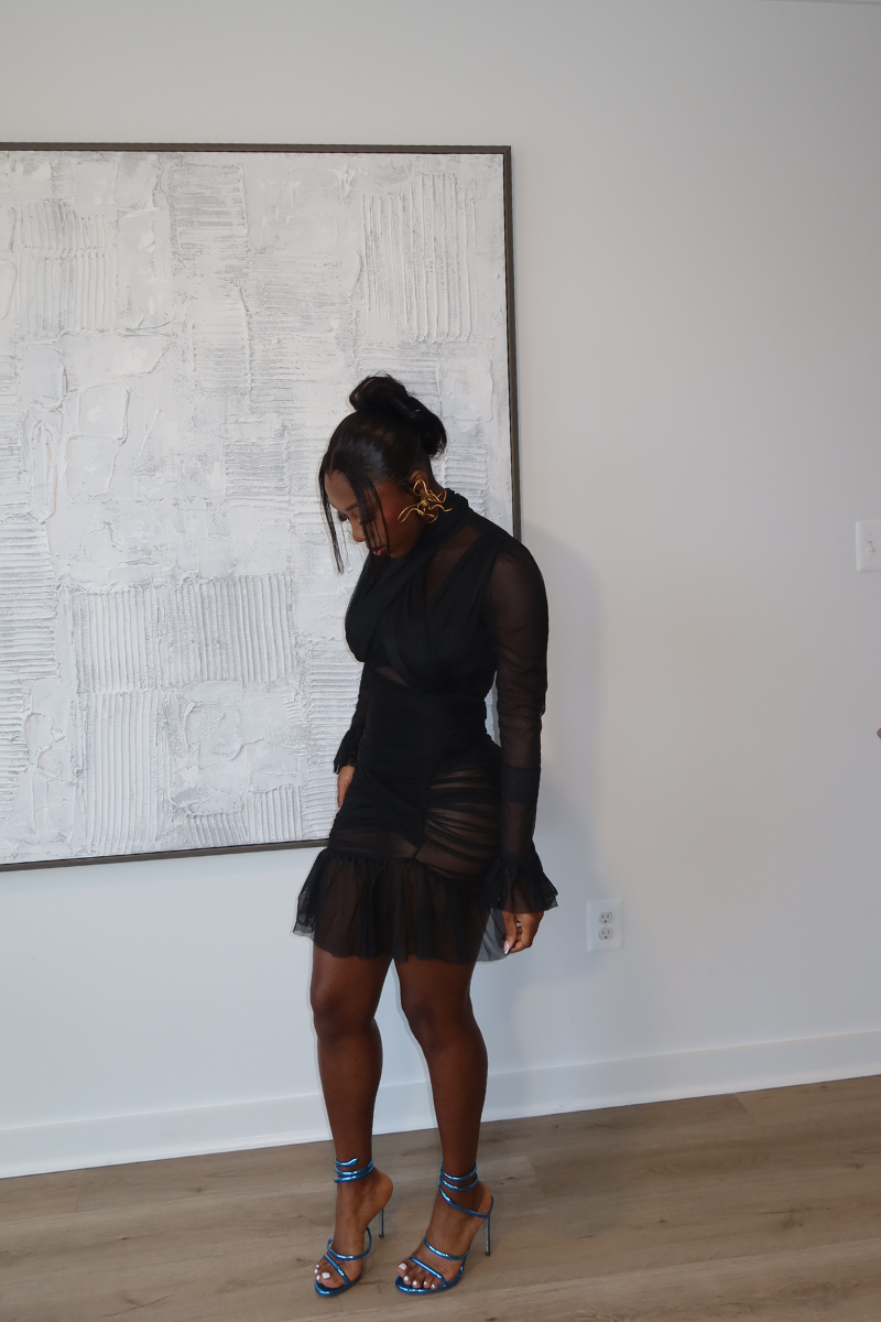 Woman wearing Demuluxe Vesper Wrap Dress, a black mesh mini with draped crisscross bodice and sheer sleeves