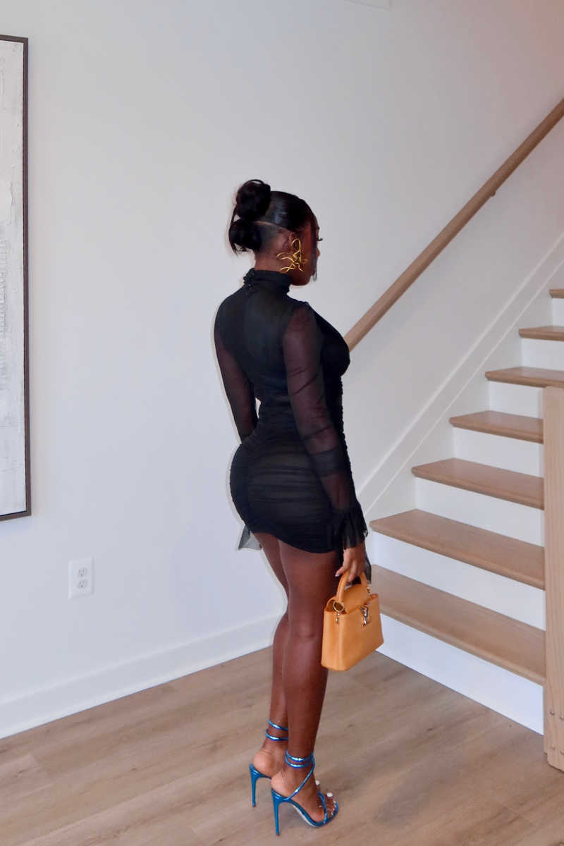 Woman wearing Demuluxe Vesper Wrap Dress, a black mesh mini with draped crisscross bodice and sheer sleeves