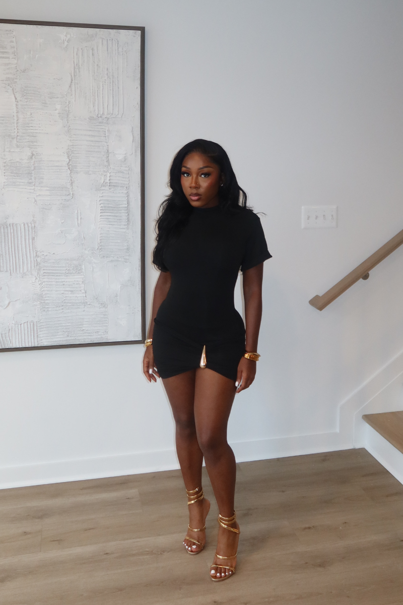Woman wearing Demuluxe Nyla Mini Dress, a black mock-neck mini dress with ruched hip detail and gold hardware