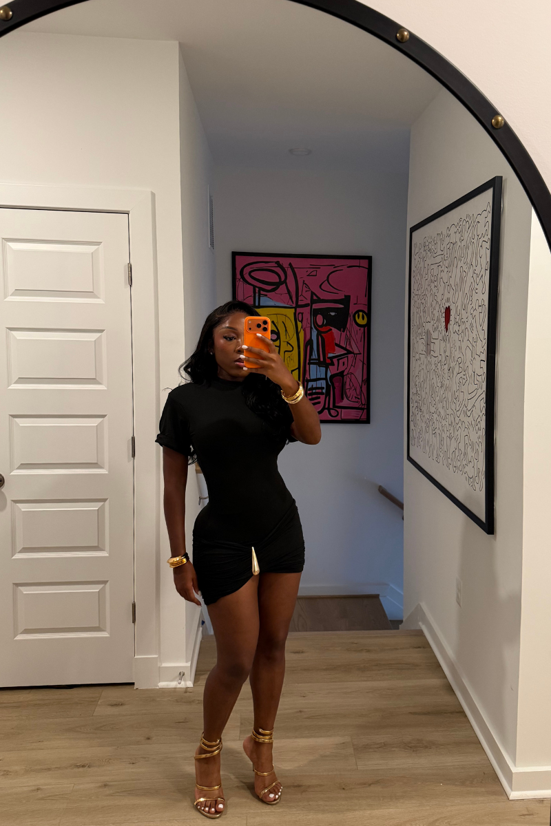 Woman wearing Demuluxe Nyla Mini Dress, a black mock-neck mini dress with ruched hip detail and gold hardware