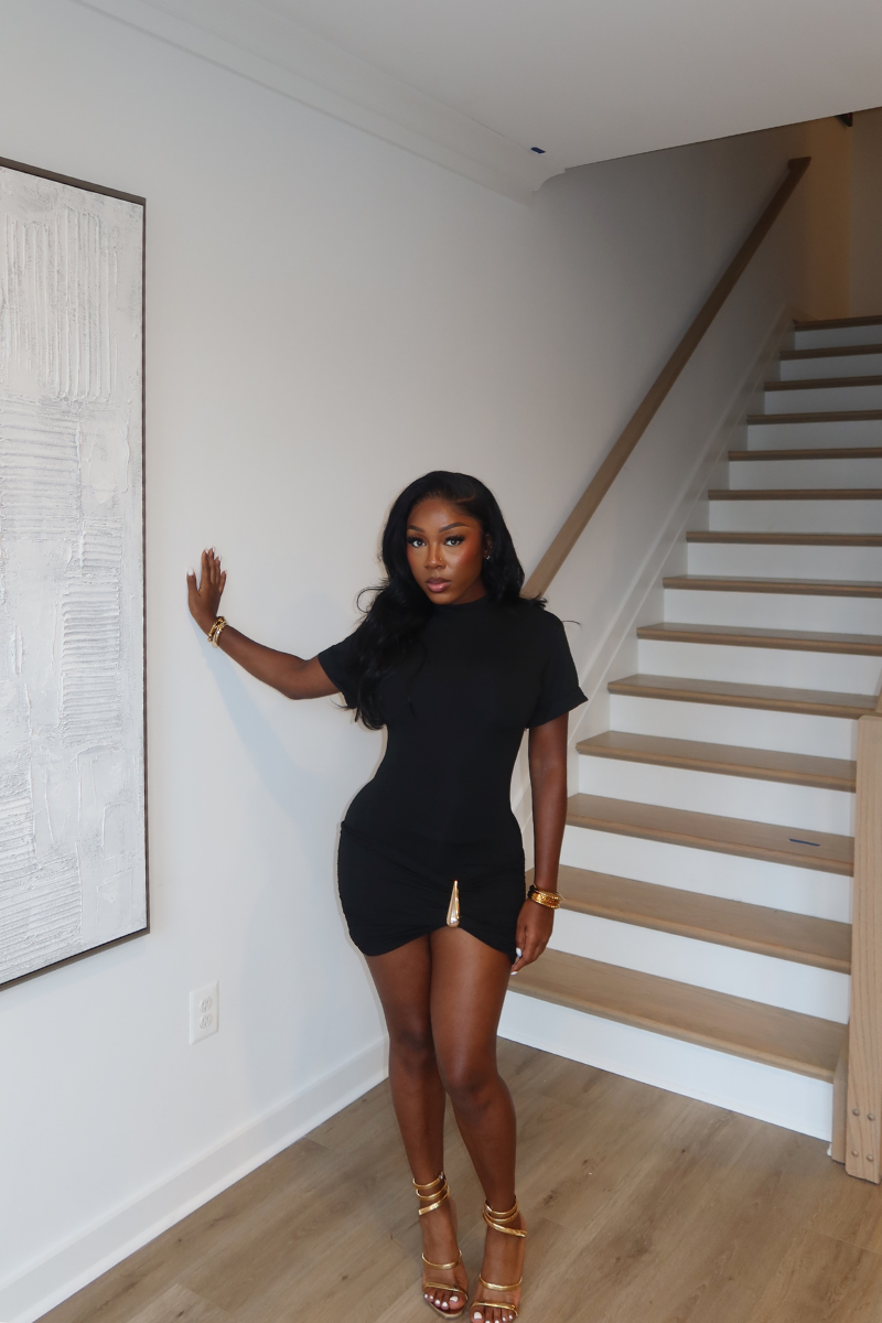 Woman wearing Demuluxe Nyla Mini Dress, a black mock-neck mini dress with ruched hip detail and gold hardware