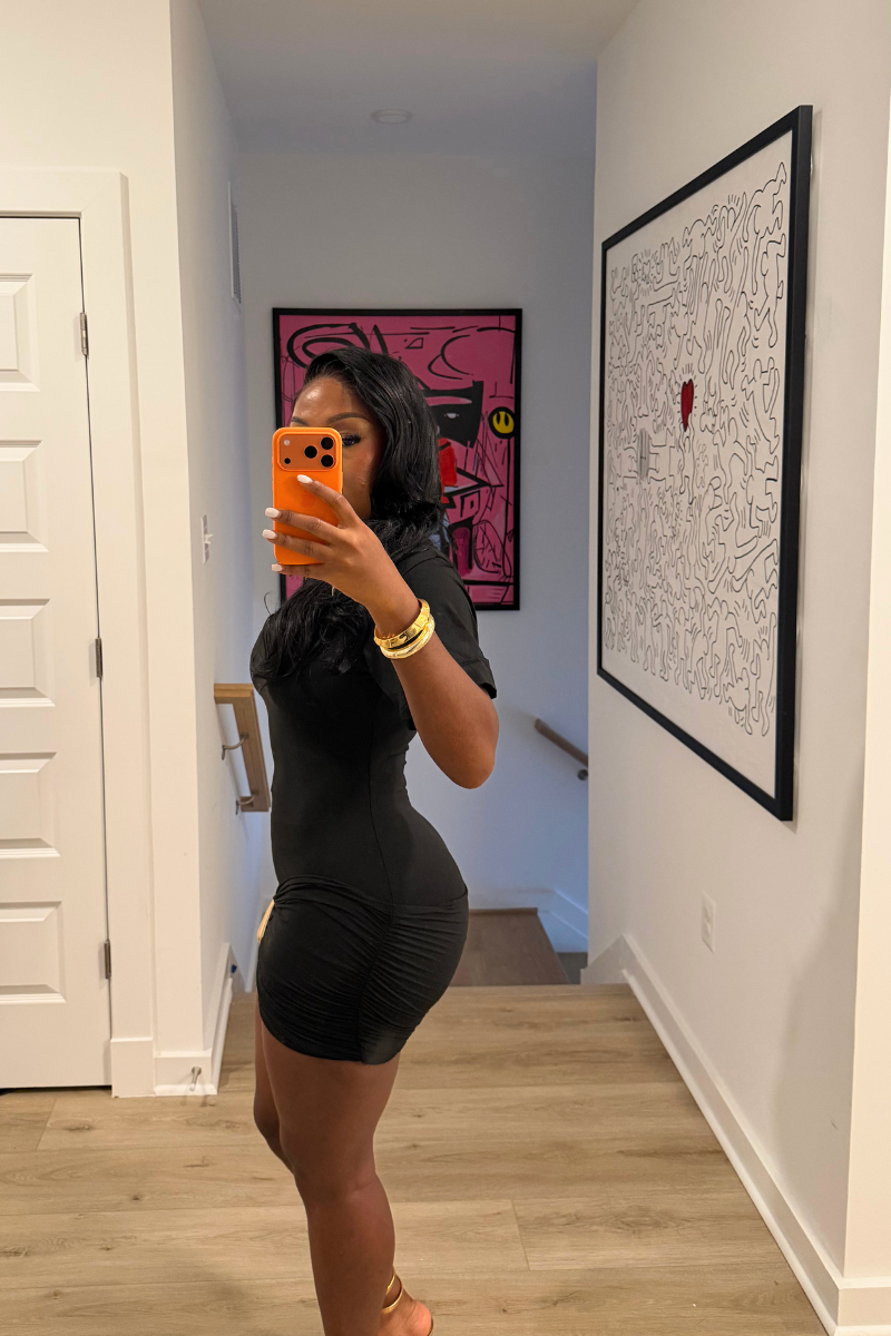 Woman wearing Demuluxe Nyla Mini Dress, a black mock-neck mini dress with ruched hip detail and gold hardware
