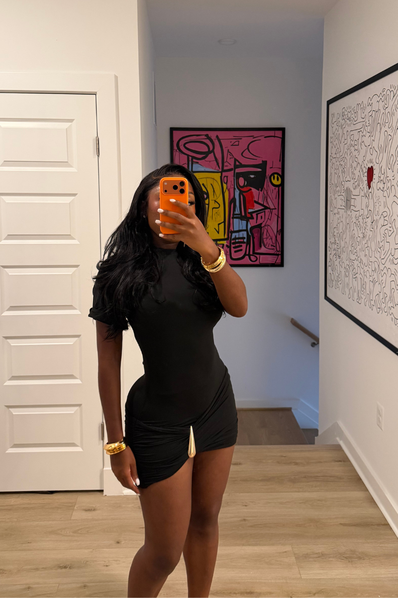 Woman wearing Demuluxe Nyla Mini Dress, a black mock-neck mini dress with ruched hip detail and gold hardware