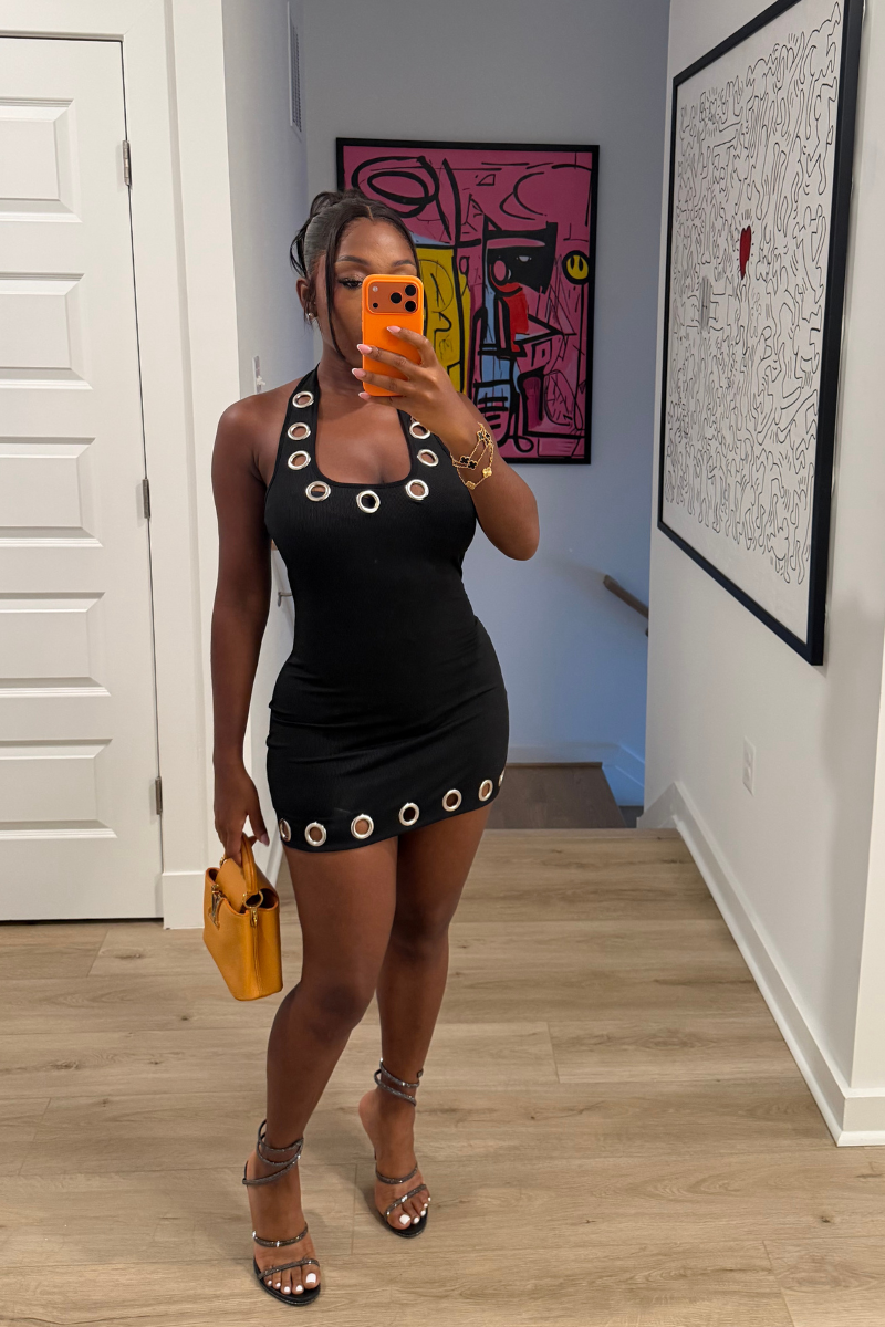 Woman wearing Demuluxe Nova Dress, a black bodycon mini with silver grommet trim at neckline and hem