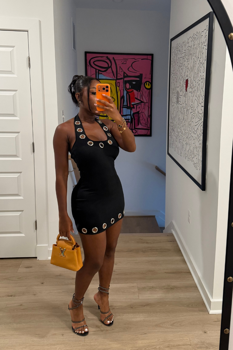 Woman wearing Demuluxe Nova Dress, a black bodycon mini with silver grommet trim at neckline and hem
