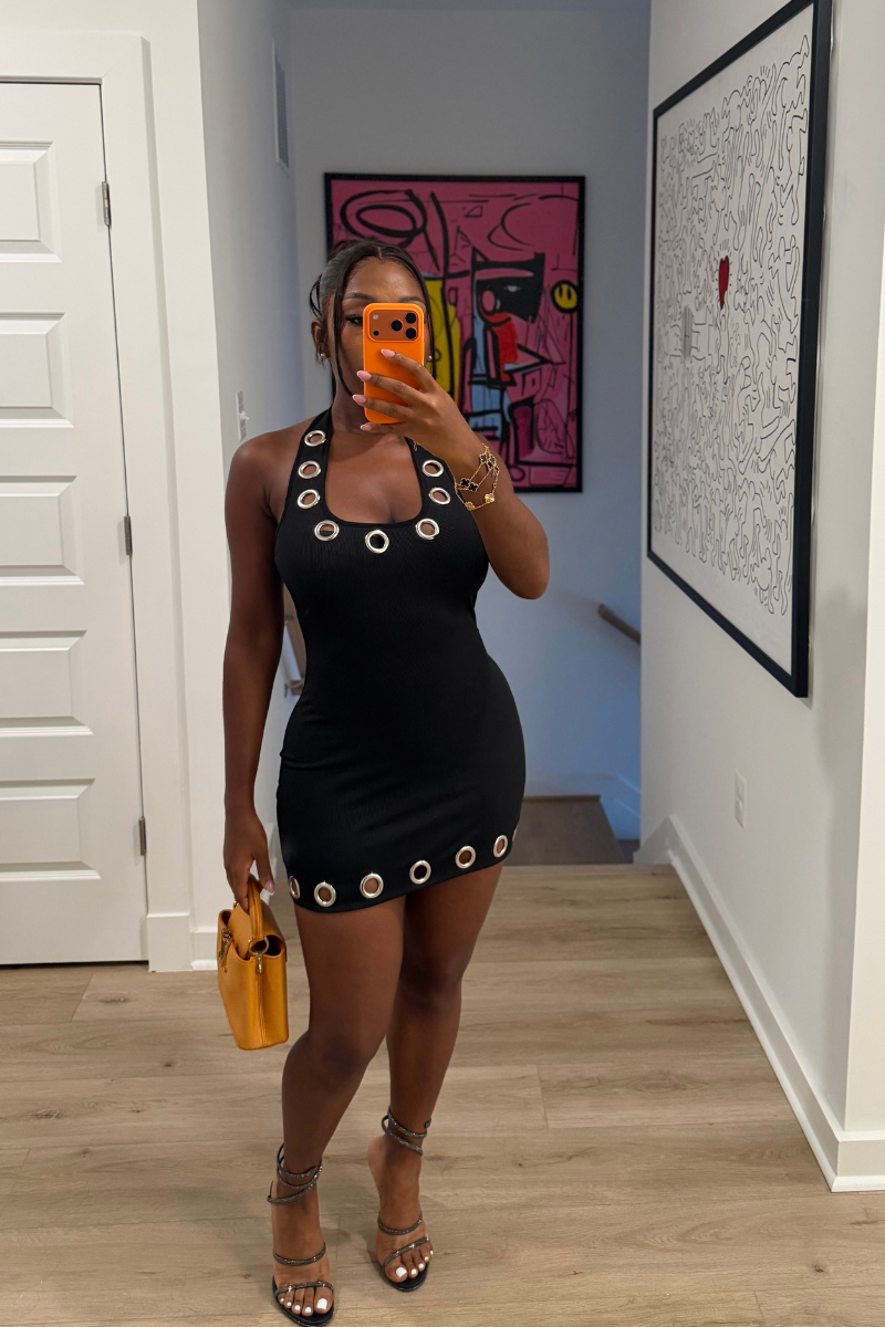 Woman wearing Demuluxe Nova Dress, a black bodycon mini with silver grommet trim at neckline and hem